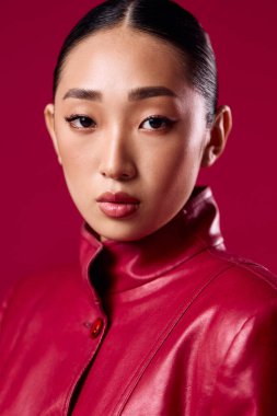 Kırmızı deri ceketli Asyalı esmer, pembe arka planda poz veriyor, kameraya ciddi bakıyor..