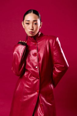 Kırmızı deri ceketli Asyalı esmer kadın elleri kalçasında kırmızı arka planda poz veriyor..
