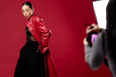 Kırmızı deri ceketli ve siyah pantolonlu Asyalı bir kadın kırmızı arka planda elinde kamerayla fotoğraf için poz veriyor..