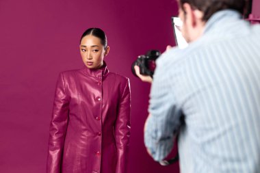 Moda Asyalı esmer. Bir adam mor deri ceketli bir kadının fotoğrafını çekiyor. Mor bir duvarın önünde duruyor..