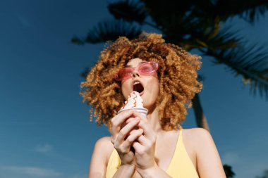 Kıvırcık saçlı neşeli bir kadın şık güneş gözlüğü takarken dondurmadan zevk alır. Titrek mavi gökyüzü ve palmiye ağaçları canlı bir yaz havası yaratır.