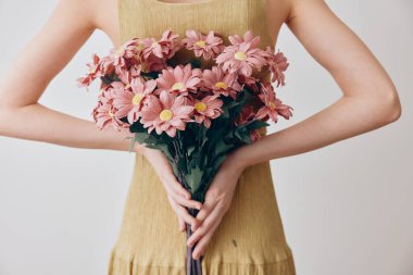 Sarı elbiseli kadın göğsünün önünde pembe papatya buketi tutuyor, elleri kalçasında..