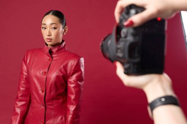 Kırmızı arka planda elinde kamerayla kırmızı deri ceketle fotoğraf çeken moda Asyalı esmer..