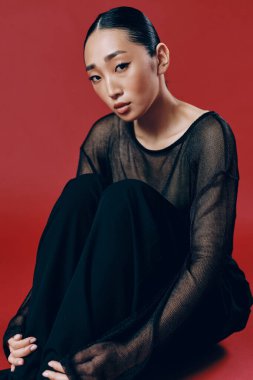 Saf bluz ve siyah pantolon giyen, elleri dizlerinin üstünde oturan ve gözleri kırmızı arka planda kapalı Asyalı bir kadın..