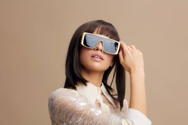 Güneş gözlüğü takan, uzun kahverengi saçlı, açık bej bir elbise giymiş, kendine güvenen ve şık bir ifadeyle tarafsız bir bej arka plana poz veren güzel bir kadın.