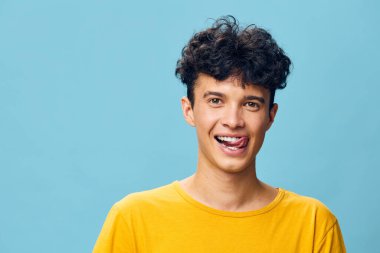 Neşeli, kıvırcık saçlı genç bir erkek, gülümserken şakacı bir şekilde dilini uzatıyor, açık mavi bir arka plana kuruluyor canlı sarı tişörtü resmin neşeli havasını arttırıyor.