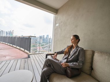 Gri takım elbiseli profesyonel bir kadın balkonda oturur ve çağdaş tasarım elementleriyle çevrili modern bir şehrin ufuk çizgisine bakar.