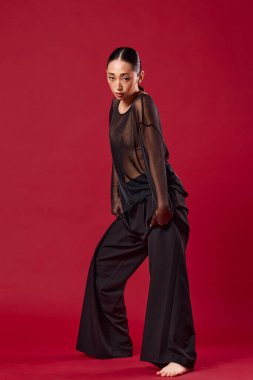 Siyah bluzlu, geniş bacaklı, elleri kalçasında kırmızı arka planda poz veren Asyalı manken..