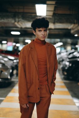 A stylish young man wearing a brown suit against the backdrop of parked cars in an indoor parking garage clipart