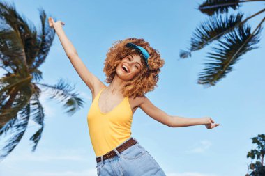 Sarı mayo ve saç bandı takan kıvırcık saçlı neşeli bir kadın, tropikal bir ortamda mutluluğu ifade ediyor.