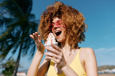 Kıvırcık saçlı ve güneş gözlüklü neşeli bir kadın dondurma tutuyor, palmiye ağaçları ve parlak mavi gökyüzü ile çevrili.