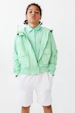 Stylish young boy in mint green jacket and white shorts strikes a pose against a modern white wall, exuding confidence and fashion-forward vibes. clipart