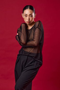 Siyah kıyafetli Asyalı manken kırmızı arka fonda poz veriyor, elleri kalçasında ve kafası yana yatık..