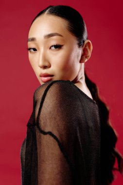 Saf siyah bluzlu Asyalı kadın elleri kalçasında kırmızı arka planda poz veriyor. Uzun saç ve modaya uygun bir tarz..