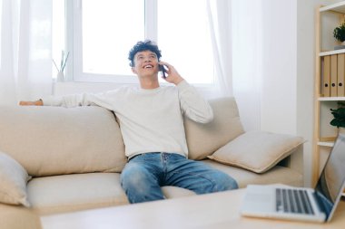 Neşeli genç bir adam parlak bir oturma odasında açık renkli bir kanepede oturuyor, bir telefon konuşmasında yer alıyor, mekan yumuşak ışıklandırma ve modern estetik, rahatlama ve rahatlama içeriyor.