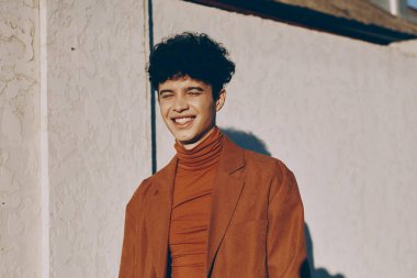 Balıkçı yaka ve ceket giyen neşeli genç bir adam, ışık duvarının önünde kendinden emin bir şekilde duruyor, pozitiflik saçıyor.