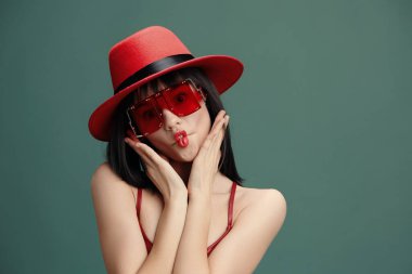 Kırmızı şapkalı, güneş gözlüklü ve kırmızı rujlu güzel Asyalı kadın yeşil arka planda poz veriyor.