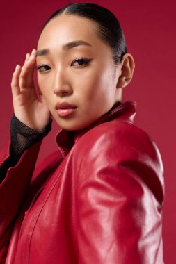 Kırmızı deri ceketli, siyah gömlekli, elleri başının üstünde kırmızı arka planda poz veren Asyalı bir esmer..