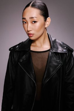 Siyah deri ceketli Asyalı kadın gri arka planda poz veriyor. Şık ve şık görünüm..