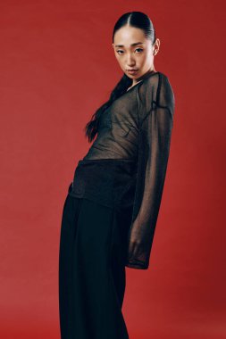 Saf bluzlu ve siyah pantolonlu moda Asyalı kadın kırmızı arka planda duruyor, elleri kalçasında ve kafası yana yatık..