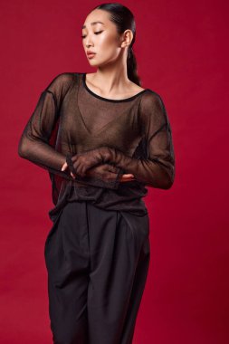 Siyah pantolonlu Asyalı bir manken ve üstü açık bir şekilde elleri kalçalarında kırmızı arka planda poz veriyor. Moda ve şık kıyafetler bir gece için mükemmel ya da özel bir etkinlik için.