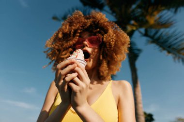 Kıvırcık saçlı genç bir kadın, güneş gözlüğü takan bir dondurma külahını palmiye ağaçlı parlak mavi bir gökyüzüne doğru tutuyor.