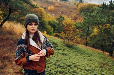 Şık bir şapkası ve ceketi olan genç bir kadın bir tepede dikilirken sonbahar ormanının renkli manzarasının tadını çıkarıyor.
