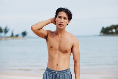 Gülümseyen Asyalı Adam Tropikal Plaj Tatilinin Tadını Çıkartıyor: Arka planda Kum ve Palmiye Ağaçları Üzerinde Kaslı Gövde