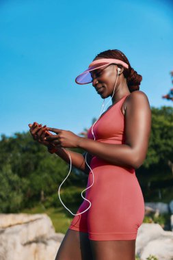 Pembe atletizm kıyafeti ve güneşliği giyen genç bir kadın açık mavi gökyüzü ve yeşil yeşile karşı telefonuyla müziğin keyfini çıkarıyor.