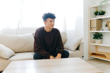 Kıvırcık saçlı genç bir adam bej renkli bir kanepede oturur, iyi aydınlatılmış bir odada, düşüncelere dalmış, yer bitkilerle ve basit mobilyalarla süslenmiştir, doğal ışıkla dolu rahatlatıcı bir atmosfer yaratır.