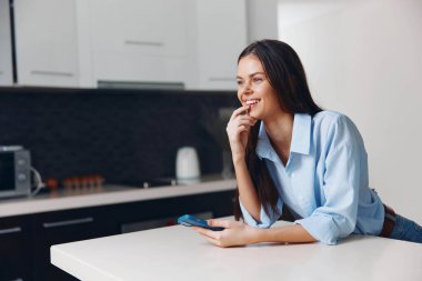 Mutfak masasında oturan mutlu kadın, elinde cep telefonuyla, neşeyle gülümsüyor ve rahatlamanın tadını çıkarıyor.