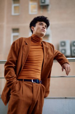 Uyumlu balıkçı yaka ve takım elbise giyen şık bir erkek model, yumuşak, nötr renkli bir binaya karşı dizilmiş.