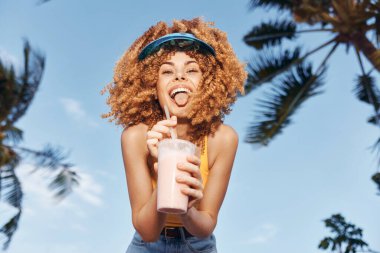 Kıvırcık saçlı, mutlu bir kadının tropikal palmiye ağaçlarının arasında berrak mavi gökyüzüne karşı smoothie yudumladığı canlı bir sahne.