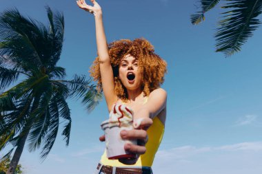 Enerjik kadın model dışarıda tropikal manzaralar arasında dondurmanın tadını çıkarırken neşe içinde gülümsüyor.