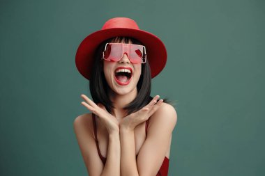 Kırmızı şapkalı ve güneş gözlüklü kadın, elleri kalçasında, geniş ağızlı komik bir yüz yapıyor, neşeli ve komik bir tutum sergiliyor, yaşam tarzı ve komedi temaları için mükemmel.