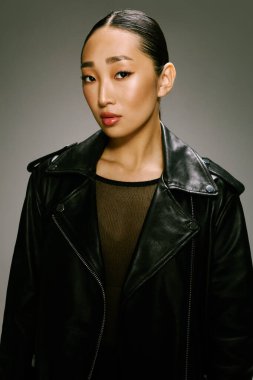 Siyah deri ceketli Asyalı kadın gri arka planda kameraya poz veriyor. Şık ve şık görünüm..