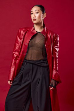 Kırmızı deri ceketli ve siyah pantolonlu Asyalı manken elleri kalçasında ve çantasında kırmızı arka planda poz veriyor..