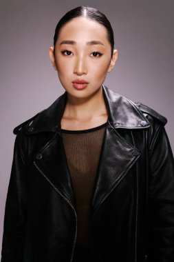 Siyah deri ceketli moda Asyalı kadın gri arka planda poz veriyor..