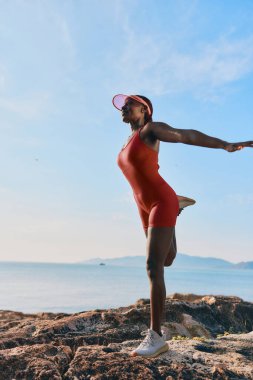 Kırmızı atletik kıyafetli bir kadın deniz kenarındaki kayaların üzerinde duruyor, yoga yapıyor gökyüzü yumuşak bulutlarla mavi