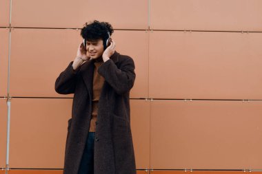 Moda Asyalı adam müzik dinliyor ve kulaklıkla turuncu duvarın önünde konuşuyor. şık ve modaya uygun görünüyor..