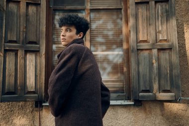 Kahverengi ceketli şık bir genç adam açık bir pencerenin önünde duruyor..