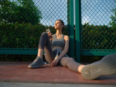 Sahada oturan genç bir kadın, elinde akıllı telefonuyla yeşillik ve çitlerle çevrili bir rahatlama anının tadını çıkarıyor.