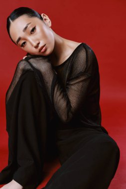 Asyalı manken kırmızı arka planda siyah pantolonlu, üstü açık, elleri dizlerinin üstünde ve başı göğsünde dinleniyor..