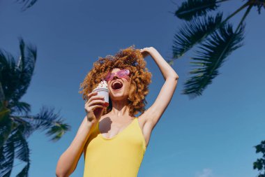 Kıvırcık saçlı ve güneş gözlüklü neşeli genç bir kadın açık mavi gökyüzüne karşı neşeyle dondurma tutuyor.