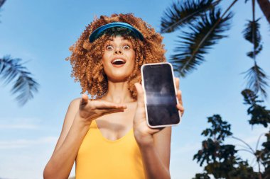Canlı bir sahne. Sarı mayo giymiş, elinde akıllı bir telefon tutan neşeli bir genç kadın. Kıvırcık saçları ve güneşliği açık havada güneşli bir günü tamamlıyor.
