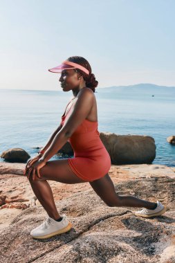 A female athlete performs a lunging stretch on rocks near the tranquil sea under bright blue skies clipart