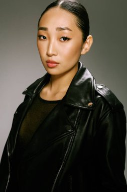 Asyalı kadın, siyah deri ceket, stüdyo resmi, gri arka plan. Genç stil sahibi Asyalı kadın, tarafsız bir arkaplana karşı deri ceketli bir fotoğraf için poz veriyor..