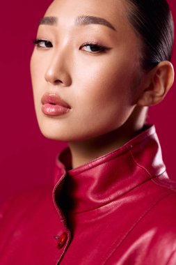 Kırmızı deri ceketli Asyalı esmer. Kırmızı arka planda poz veriyor. Gözleri kapalı ve dudaklarında kırmızı ruj izi var..