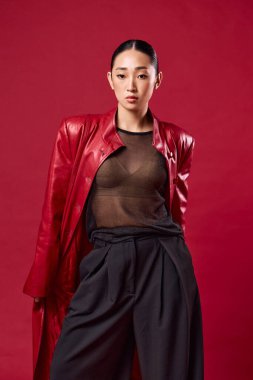 Kırmızı deri ceketli, siyah pantolonlu, elleri kalçasında kırmızı arka planda poz veren Asyalı bir kadın. şık ve şık görünüyor..