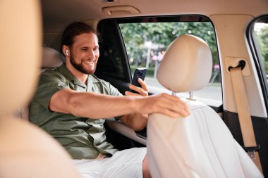 Modern şehir yaşam tarzına odaklanmış, akıllı telefon ve kulaklıklarını takmış, bir arabada oturan mutlu genç bir adam.